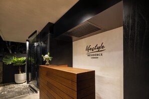 A reception desk with a wooden finish, a potted plant, and a wall with the text 'lifestyle residence CANGGU'. - Body Factory Lifestyle Residence Canggu (Canggu)