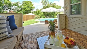 Un patio con una mesa de madera, una silla con cojín y una bandeja blanca con piña, limonada y fruta.