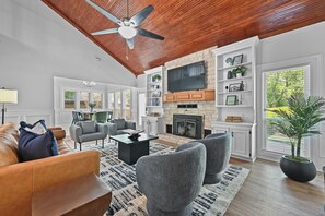 A spacious living room with a fireplace, a ceiling fan, and a large TV mounted above it.