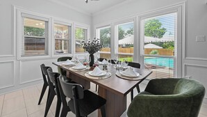Comedor con una larga mesa dispuesta para seis comensales, paredes blancas con paneles, ventanas amplias con persianas y una araña.