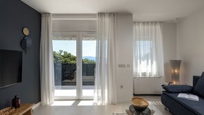 Una sala de estar moderna con una pared oscura, un televisor de pantalla plana, un sofá, una mesa de centro y un amplio ventanal con cortinas.
