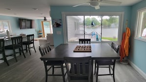 A dining area with a large table and chairs, a TV mounted on the wall, and a view of a lake through large windows. - Newly Renovated, Lakefront, Dog Friendly Cottage, Sand Lake, Harrison (Harrison)