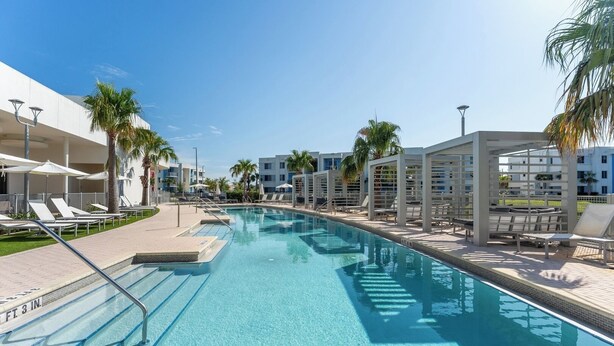 A swimming pool with lounge chairs and umbrellas, surrounded by palm trees and modern buildings. - Daytona 2BR Getaway Parking Family & Pet-friendly (Daytona Beach)