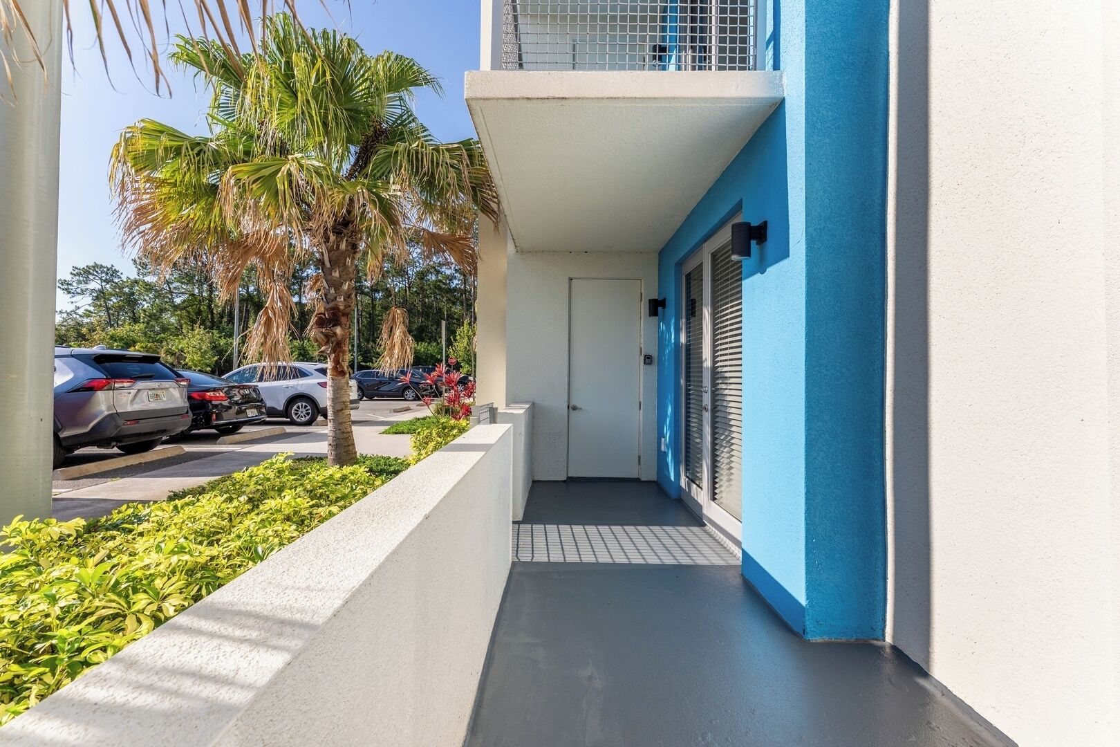 A narrow pathway leading to a building with a blue and white exterior, a door, and a small balcony with a metal railing.