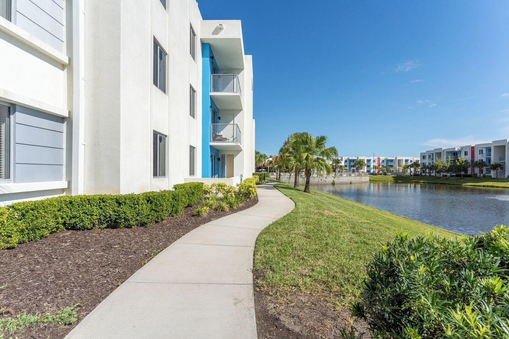 A modern hotel with a white facade, a blue accent on the second floor, and a pathway leading to a body of water.