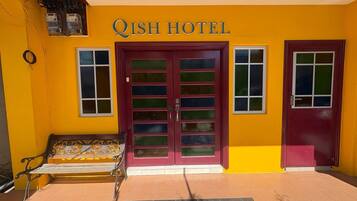 Entrada de un hotel con fachada amarilla, puerta roja y un banco afuera.
