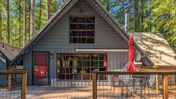 A cabin with a large window and a red door, surrounded by a deck with chairs and a table. - Charming, rustic cabin on the golf course (Packwood)