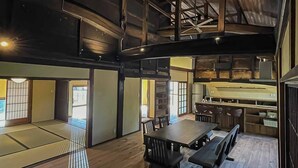 A spacious interior with a high ceiling, wooden beams, a dining area with a long table and chairs, and a kitchen area with a counter and stools.