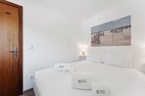 A hotel room with a wooden door, a bed with white bedding, two pillows, and a nightstand with a lamp. There is a wall-mounted artwork depicting a beach scene. - GuestReady – Ideal Couple’s Spot in Gaia (Vila Nova de Gaia)