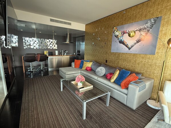 Een moderne woonkamer met een grijze bank, kleurrijke kussens, een glazen salontafel en een groot abstract schilderij aan de muur.