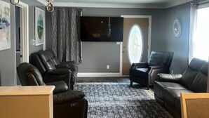 A modern living room with dark wood flooring, a large black leather sofa, a smaller sofa, and a recliner. There is a flat-screen TV mounted on the wall, a large window with curtains, and a patterned rug. - Spacious 4-bedroom fully remodeled home in charming Sandusky (Sandusky)