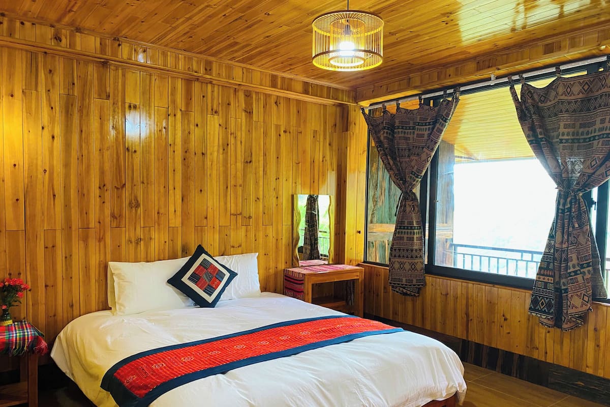 A wooden-paneled room with a bed, a small table, and a window with curtains.