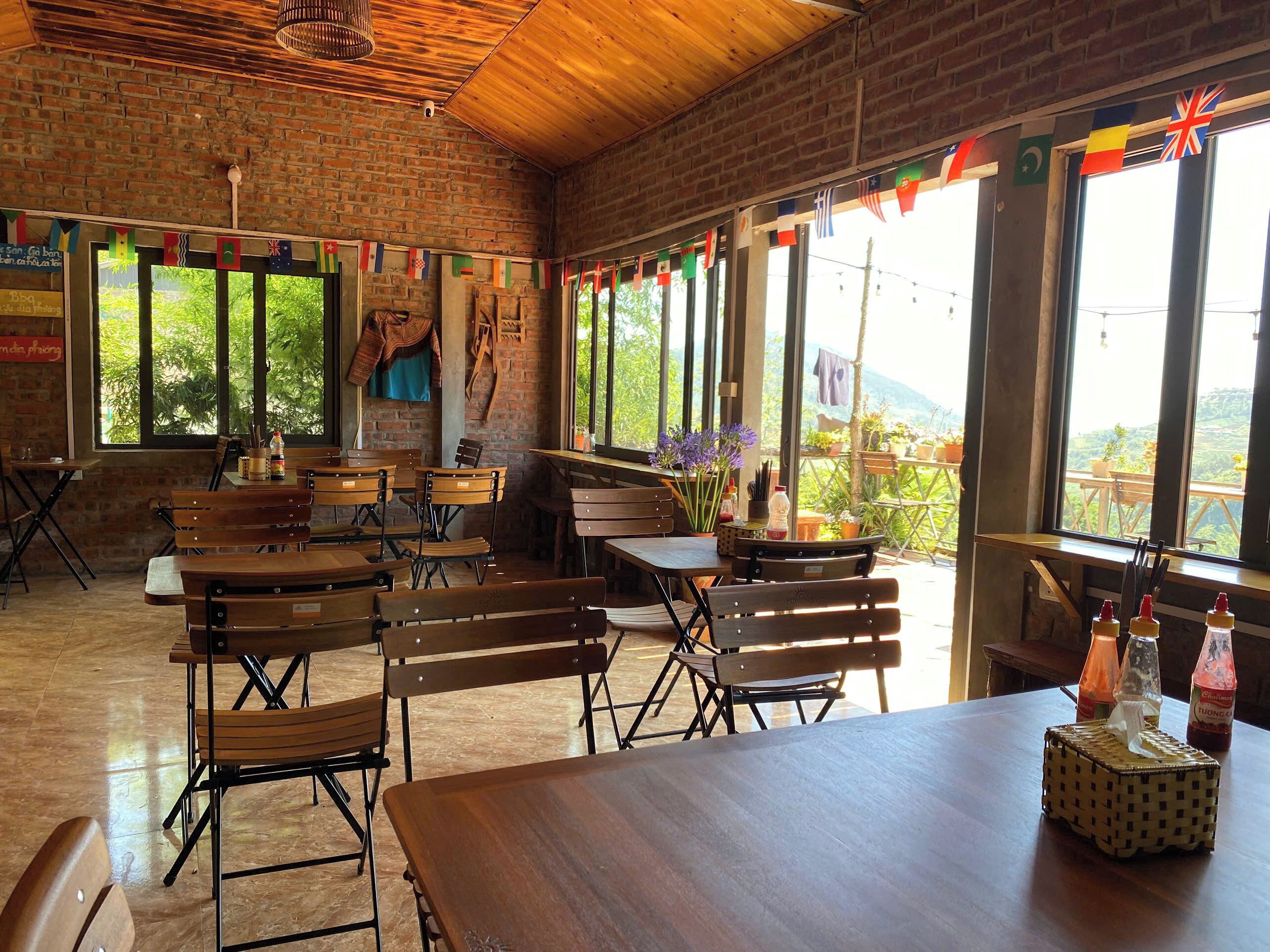 Un intérieur rustique avec des murs en briques, des tables et des bancs en bois, de grandes fenêtres ornées de drapeaux et une vue sur la verdure à l’extérieur.