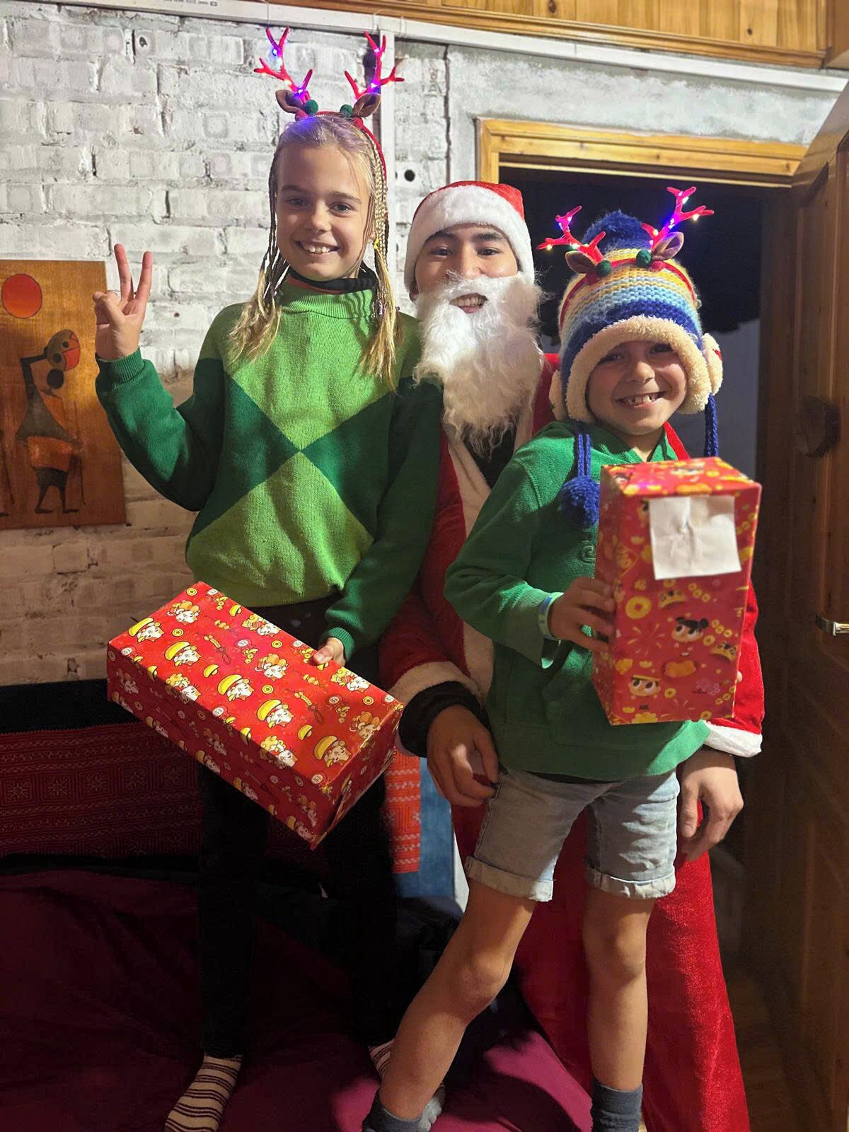 Une pièce avec un mur en briques, une porte en bois et un tableau. On y voit trois personnes, dont une porte un bonnet de Père Noël et tient une boîte cadeau.