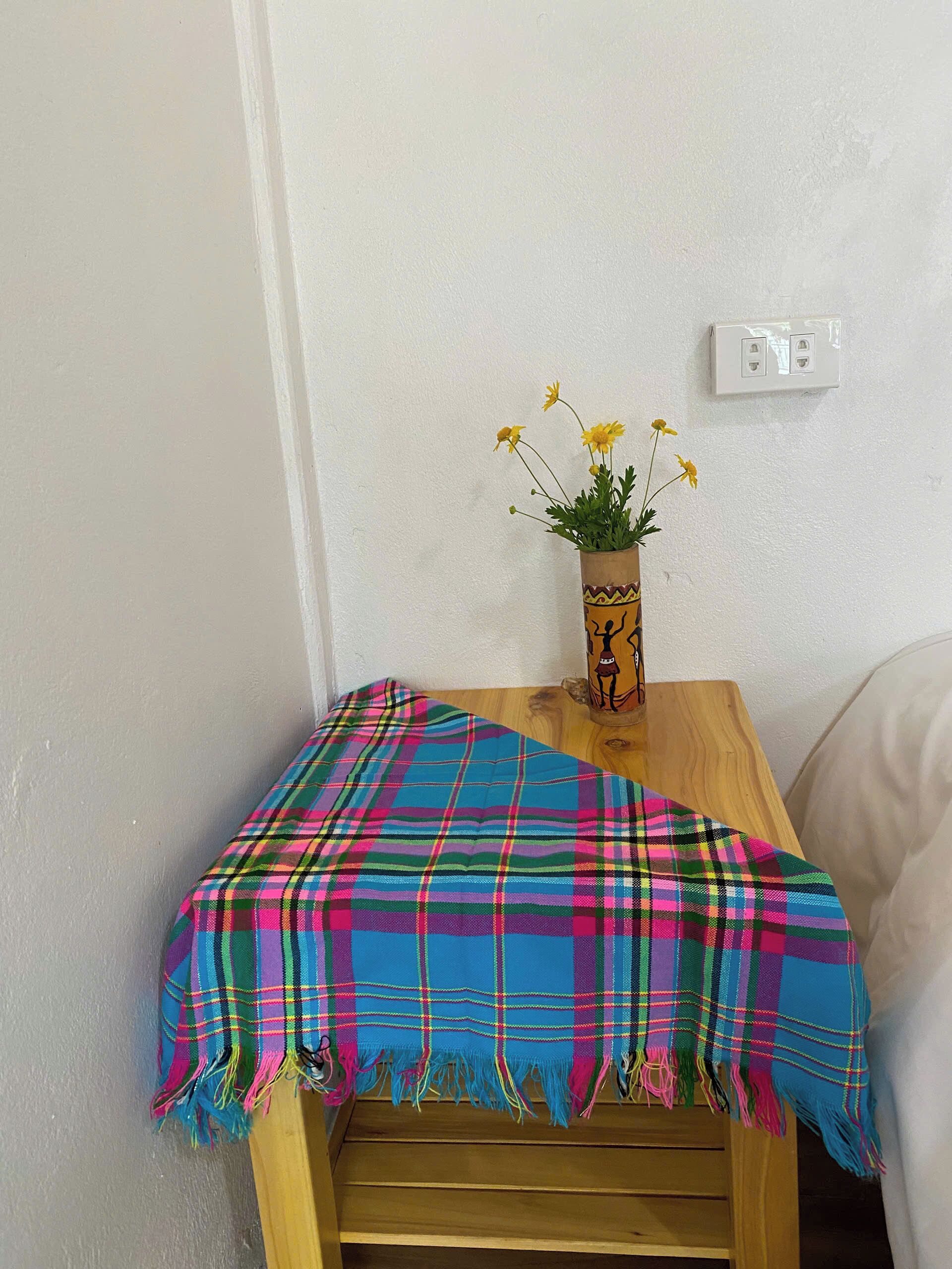 Un banco de madera con una manta colorida de cuadros, un jarrón con flores amarillas y una pared blanca con un interruptor de luz.