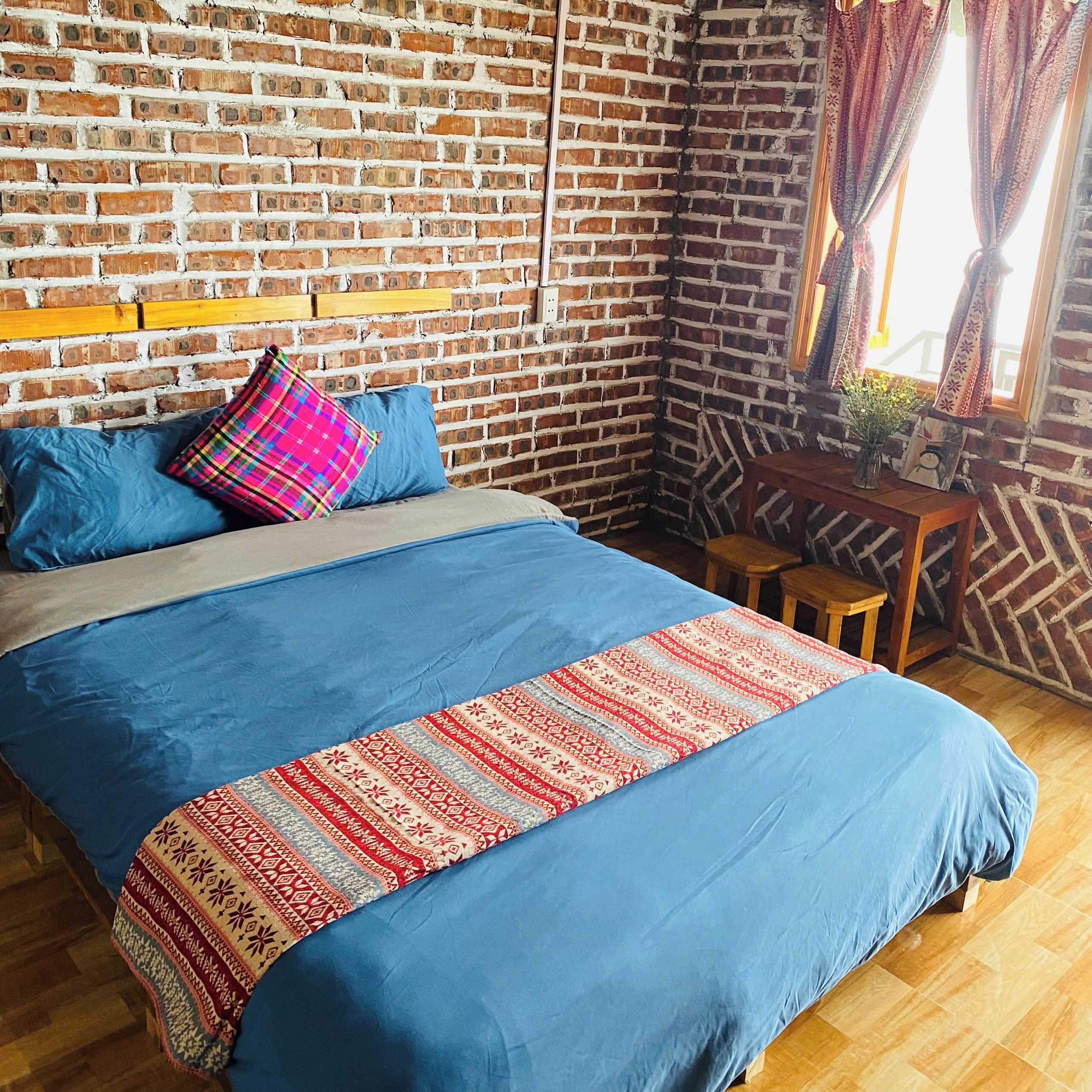 A room with a brick wall, a bed with blue bedding and a patterned throw, a wooden table, and a window with curtains.