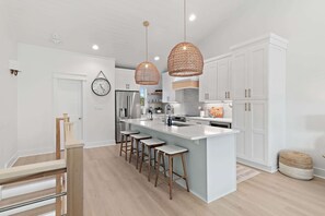 Une cuisine moderne avec des meubles blancs, un îlot central doté de tabourets, des luminaires suspendus et un réfrigérateur en acier inoxydable.
