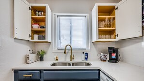 Een keuken met een dubbele spoelbak, witte kastjes, een raam met jaloezieën en een koffiezetapparaat.