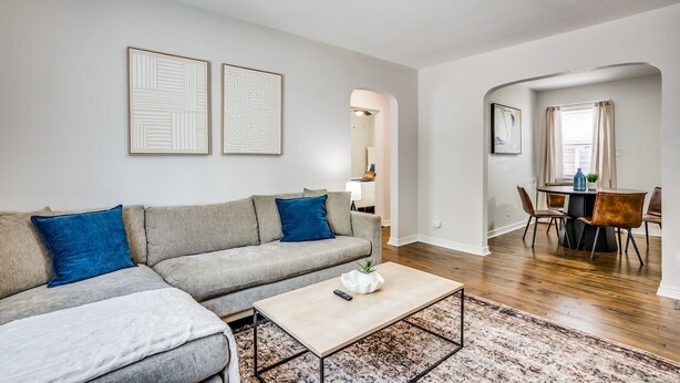 Een woonkamer met een grijze bank, blauwe kussens, een salontafel en een vloerkleed. Er is een eethoek met een tafel en stoelen.
