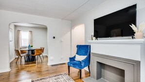 Een woonkamer met een open haard, een blauwe fauteuil, een eethoek met een ronde tafel en stoelen, en een televisie boven de open haard.