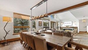 Un comedor amplio con una mesa y sillas de madera, una lámpara colgante moderna, y vistas de una sala de estar acogedora con chimenea.