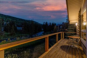 A wooden deck with a table and chairs, overlooking a forested valley and a river. - Vail Village Riverfront Retreat Hot Tub, In Town (Vail)