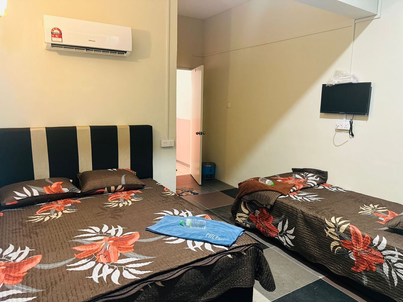 A hotel room with two single beds, an air conditioner, a television, and a small table with a water bottle.