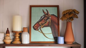 A framed horse painting, a tall white candle, a vase with dried flowers, a wooden sculpture, and a ceramic mug on a wooden shelf.
