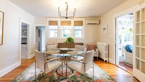 A dining area with a glass table, four chairs, a chandelier, a radiator, and a view through a window.
