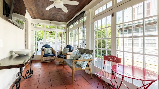 A sunlit room with a ceiling fan, wicker furniture, a red metal table, and large windows.