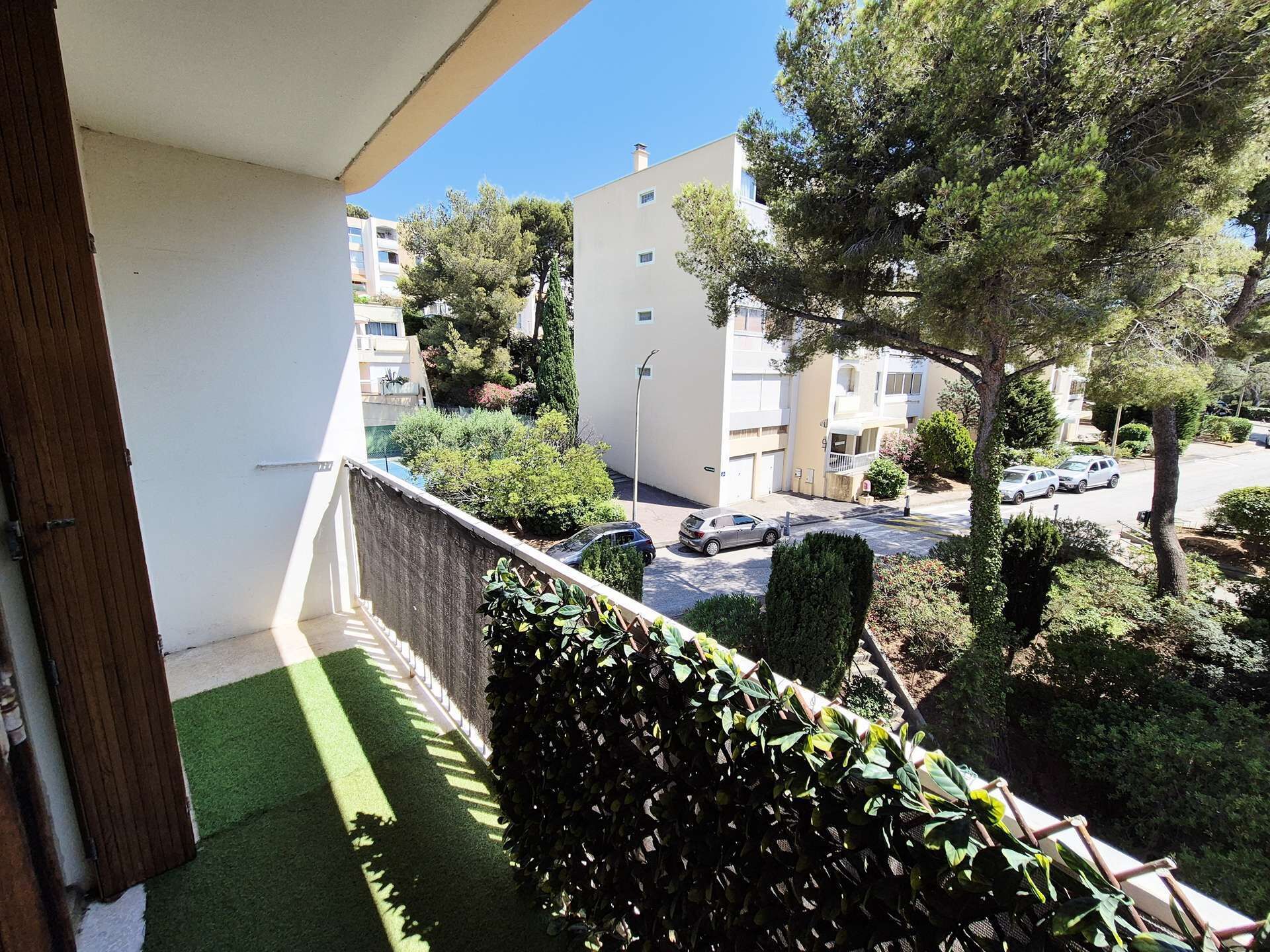 A balcony with artificial grass, a hedge, and a view of a residential street with parked cars and buildings.