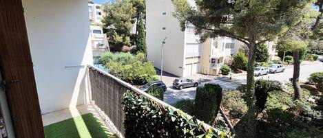 A balcony with artificial grass, a hedge, and a view of a residential street with parked cars and buildings.