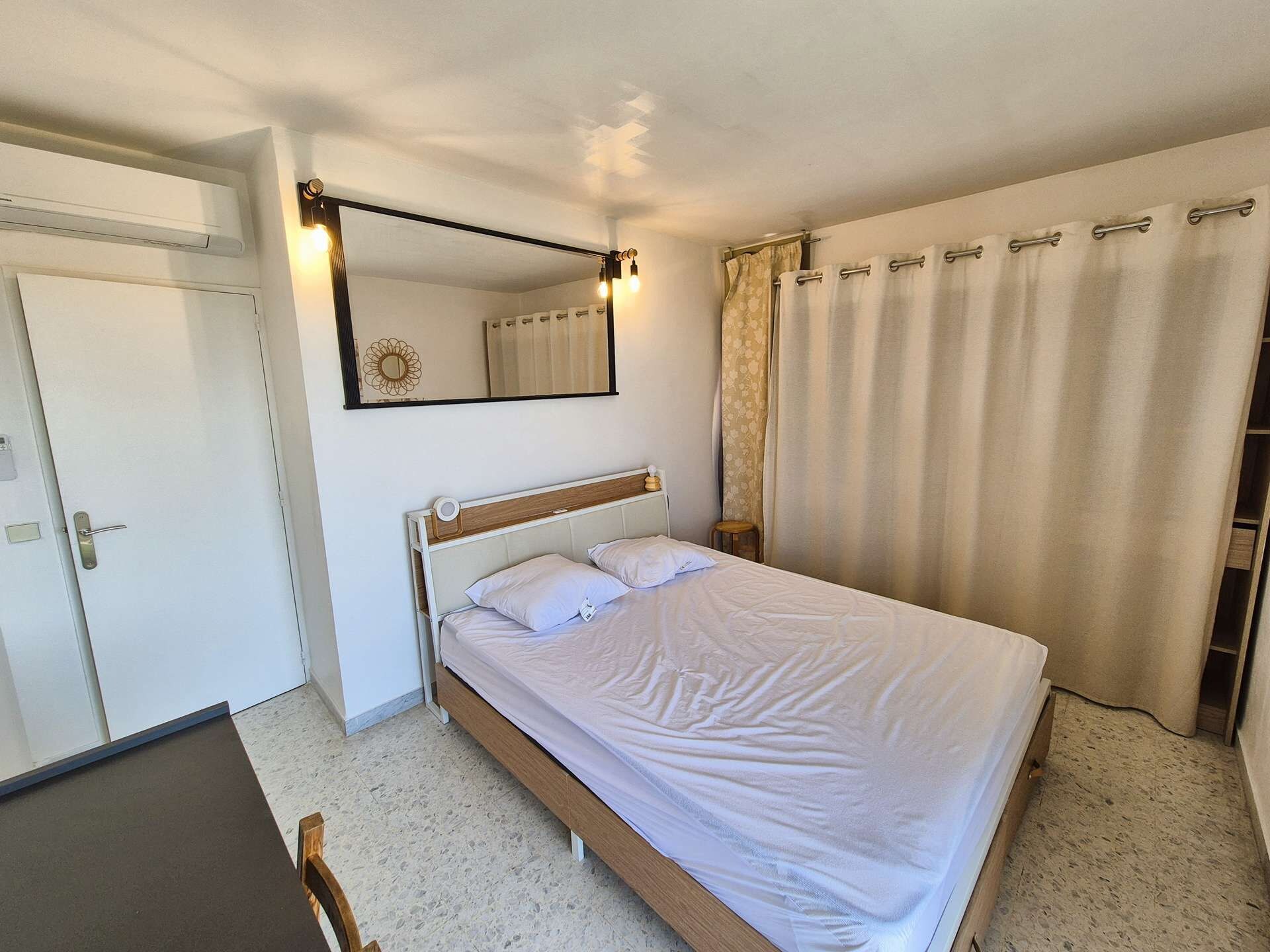 A bedroom with a bed, a bedside table, a mirror, and a curtain.
