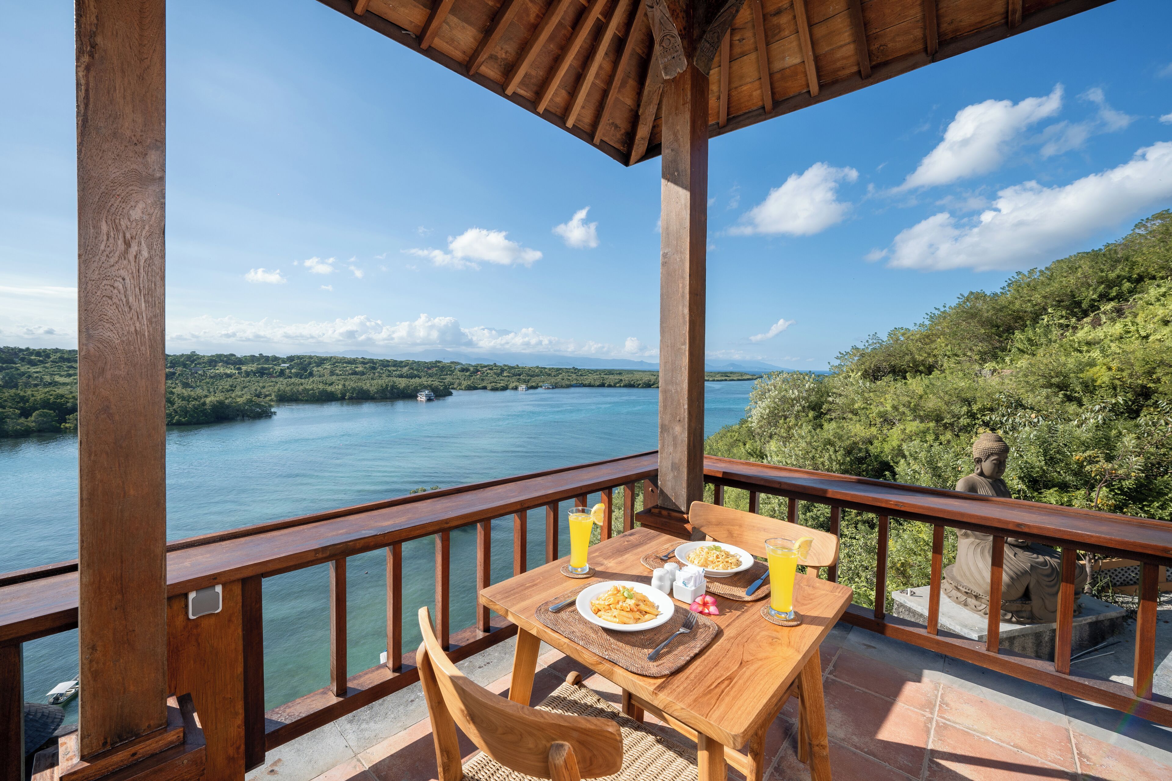 A wooden deck with a table set for two, overlooking a lake with a Buddha statue in the background.