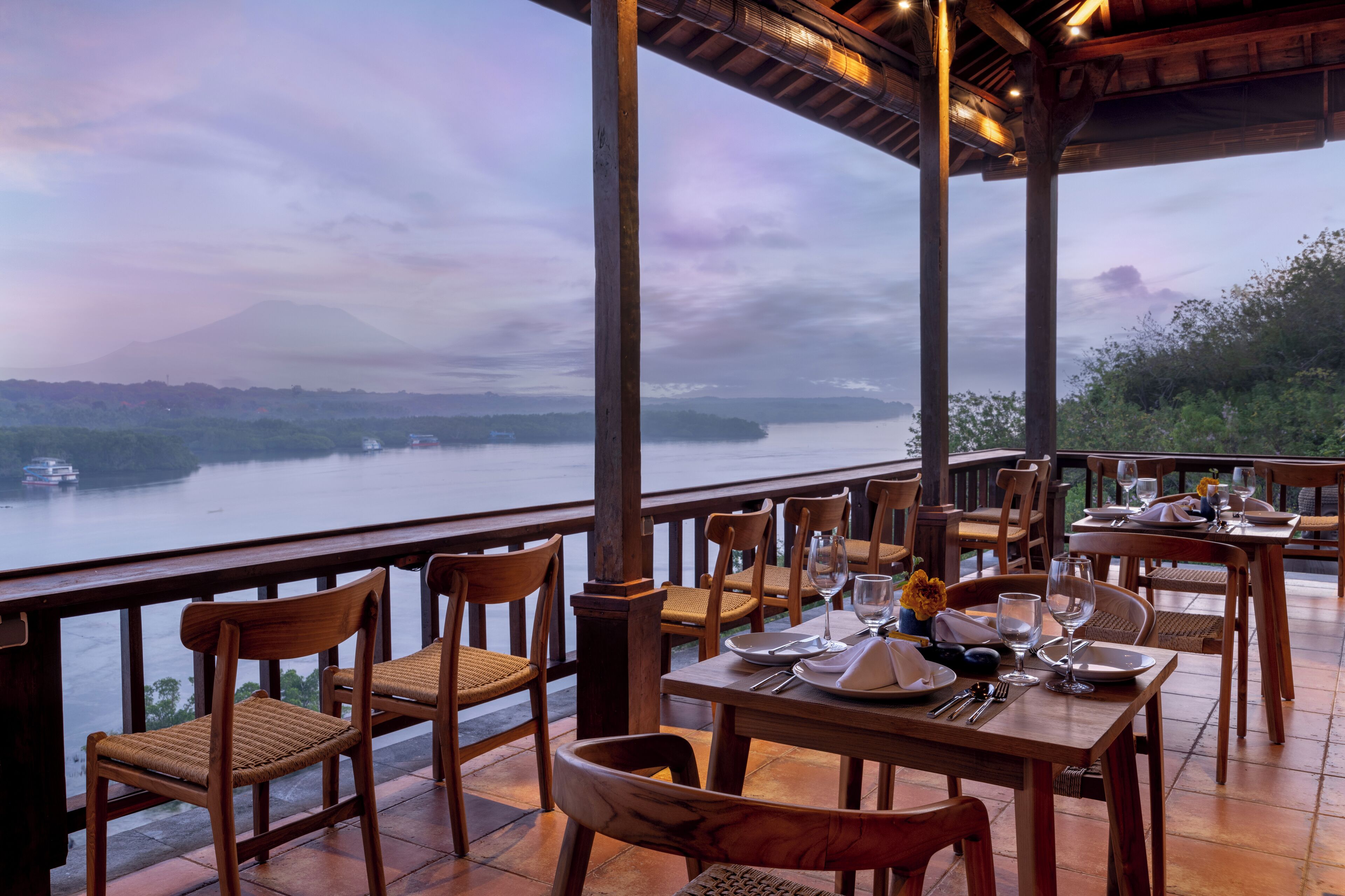 A restaurant with wooden furniture and a table set for dining, overlooking a body of water and a mountain.