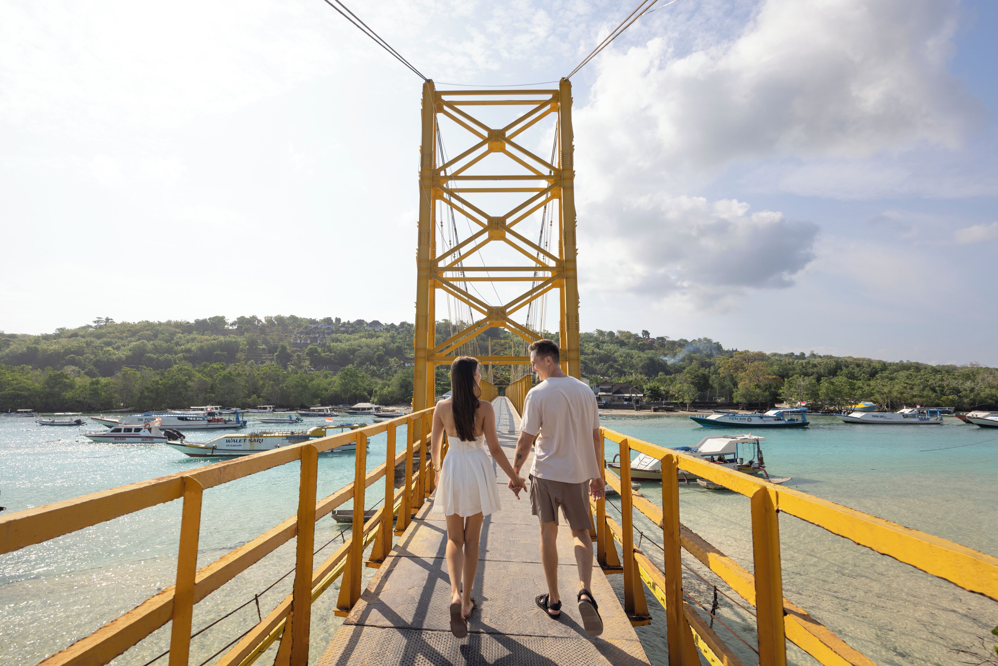 A couple walking hand in hand on a yellow bridge with boats in the background.