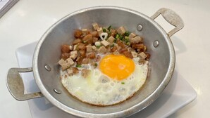 En metallpanna med ett stekt ägg och tärnat kött på en vit yta.