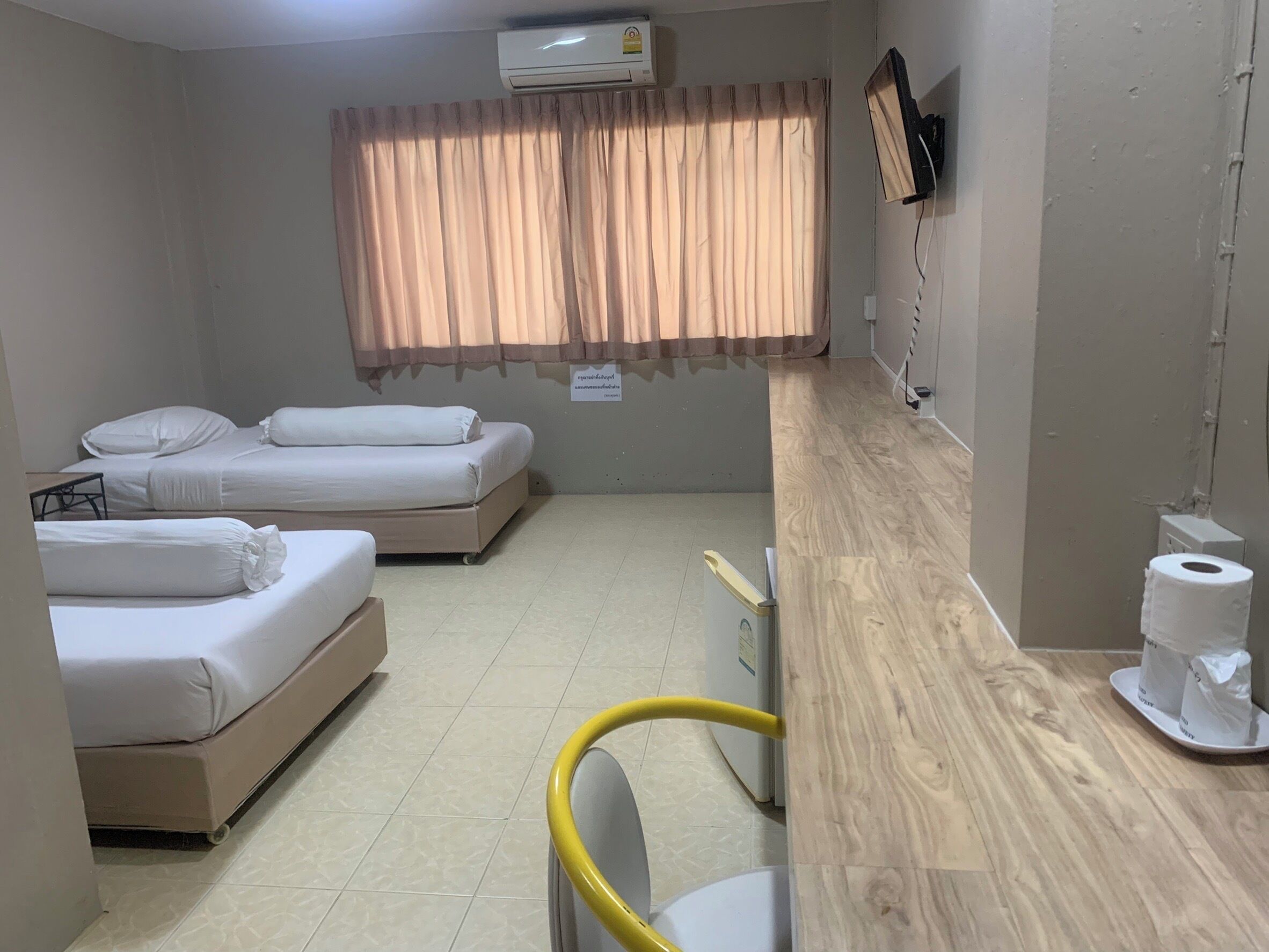 Habitación de hotel con dos camas, un escritorio de madera, una silla, un equipo de aire acondicionado y una ventana con cortinas.