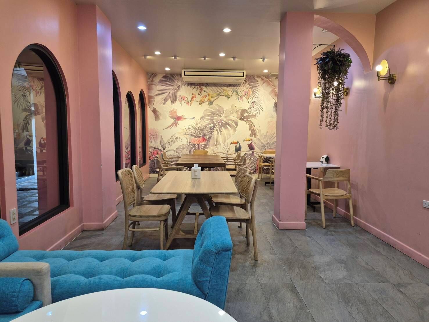 A dining area with a long table and chairs, a blue sofa, and a mural with birds and plants on the wall.