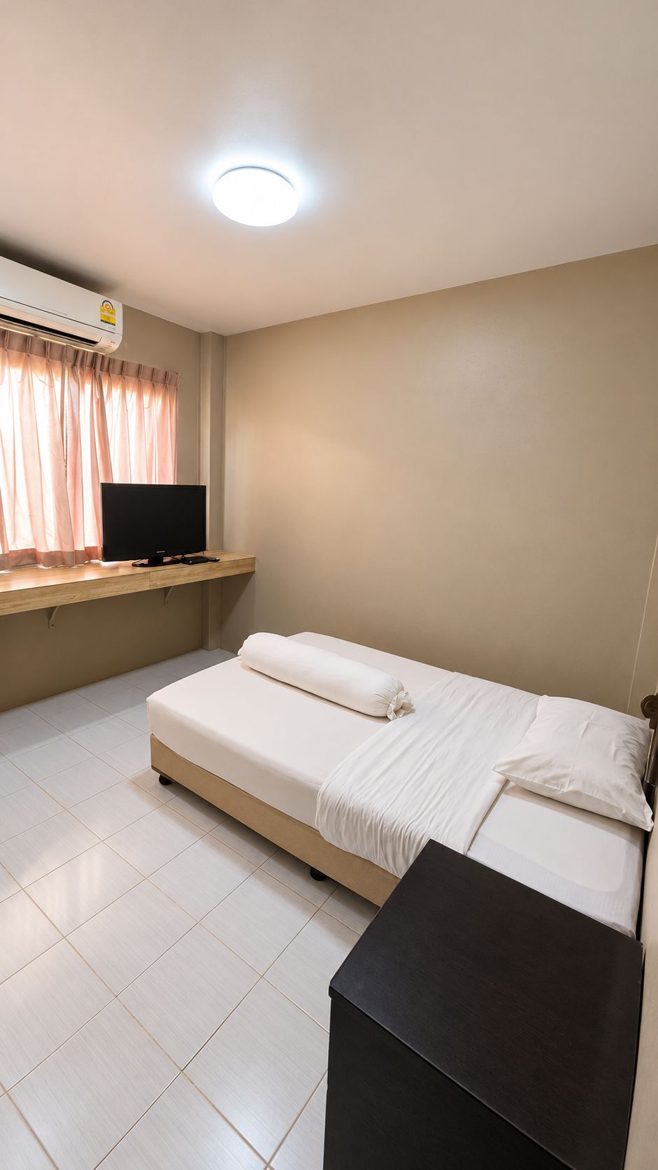 Una cama individual con ropa de cama blanca, un televisor de pantalla plana sobre un estante de madera y un aire acondicionado instalado en el techo.