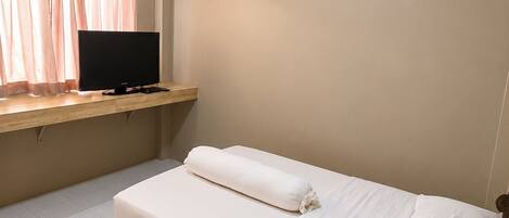 Una cama individual con ropa de cama blanca, un televisor de pantalla plana sobre un estante de madera y un aire acondicionado instalado en el techo.