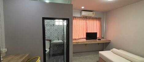 A compact room with a bed, a desk with a computer, a bathroom with patterned tiles, and a window with curtains.