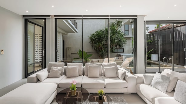 Ein modernes Wohnzimmer mit einem großen Ecksofa, einem Glastisch und Blick auf einen Balkon mit Pflanzen.