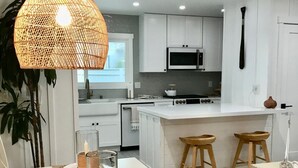 Une cuisine moderne avec des meubles blancs, un luminaire suspendu et un coin repas agrémenté d’assiettes, de serviettes et d’un panier.