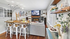 Un salon moderne avec une cheminée, un coin repas avec des chaises blanches et un téléviseur fixé au mur.