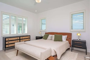 A bedroom with a large bed, two bedside tables, a rattan console, and a window with blinds. - Oceans 9 LUXE | 4BR Beachfront Villa in West Bay (West Bay)