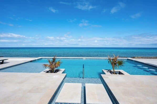 A rectangular infinity pool with clear blue water, surrounded by a light-colored tiled deck. - Oceans 9 LUXE | 4BR Beachfront Villa in West Bay (West Bay)