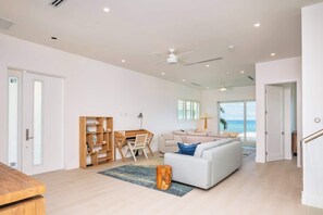 A spacious living area with a white sofa, a wooden bookshelf, a dining table, and a view of the ocean. - Oceans 9 LUXE | 4BR Beachfront Villa in West Bay (West Bay)