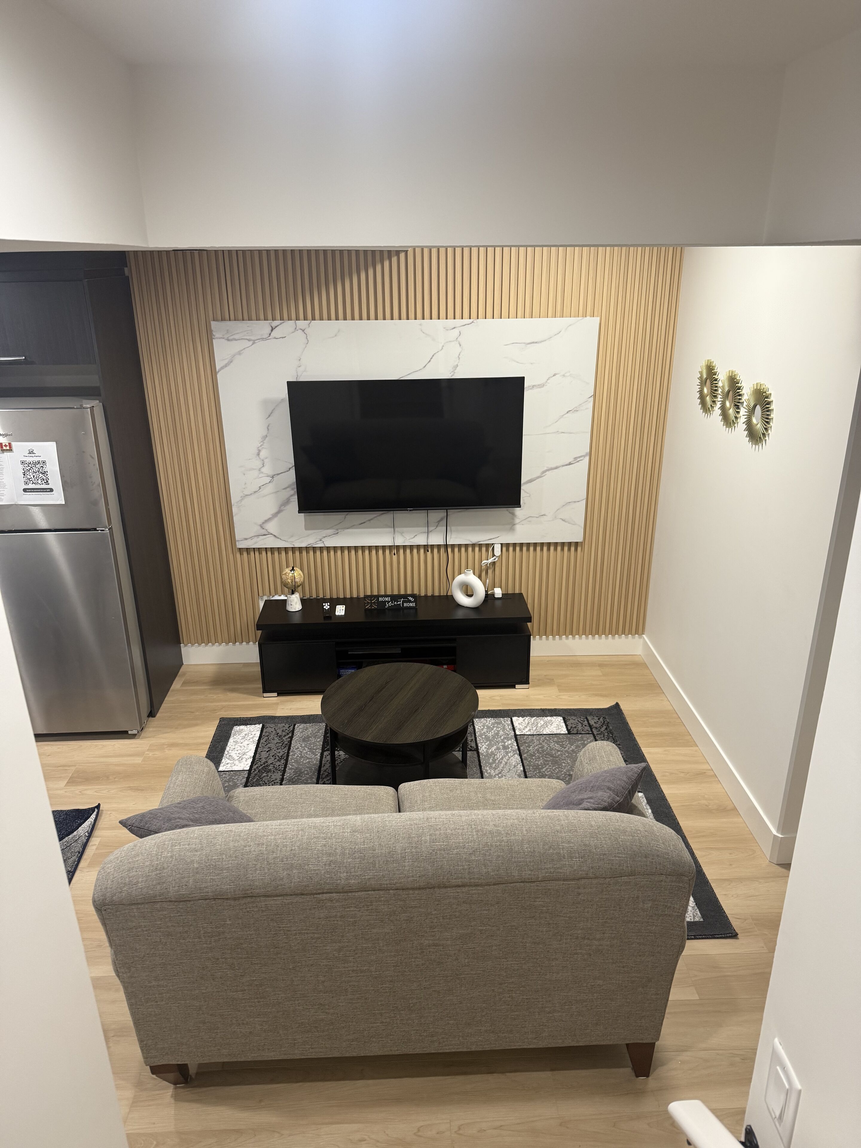 Ein modernes Wohnzimmer mit einem wandmontierten Flachbildfernseher auf einer Wand mit Marmormuster, einer schwarzen Unterhaltungsecke darunter, einem runden Couchtisch und einem grauen Sofa.