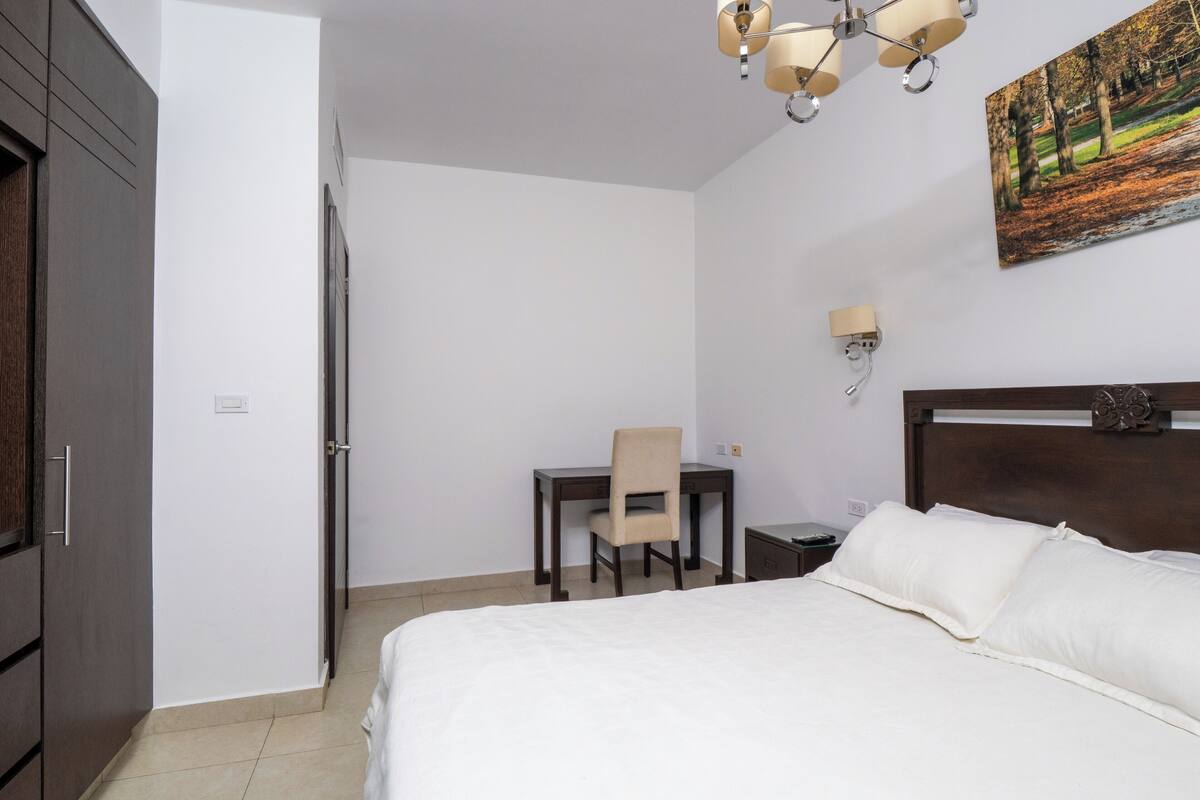 A hotel room with a bed, a desk, a chair, a nightstand, a lamp, and a painting on the wall.